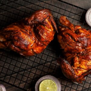 Juicy grilled chicken halves on a cooling rack, garnished with sliced lemon and red onions.